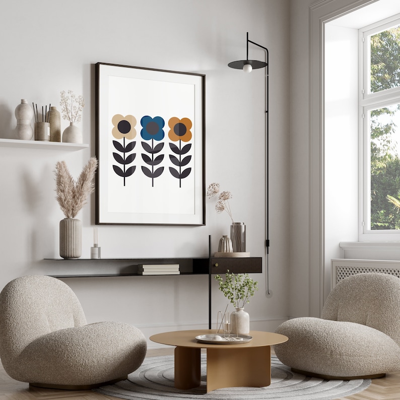 Op de afbeelding: Een ingelijste print van drie gestileerde bloemen met een witte achtergrond. De bloemen zijn geel, blauw en oranje met zwarte stelen en bladeren. De print hangt aan een witte muur boven een zwarte plank.