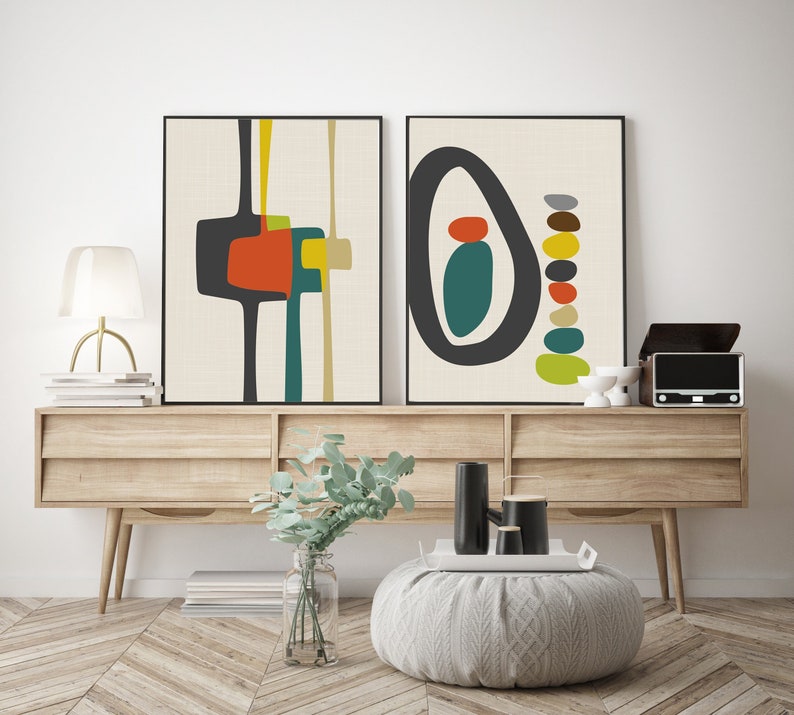 May include: Two framed wall art prints featuring abstract geometric shapes in black, orange, yellow, and teal on a cream background. The left print displays vertical lines and block forms, while the right print shows a large oval and stacked pebbles.