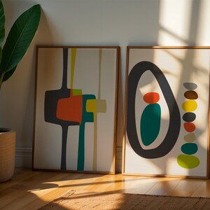 May include: Two framed rectangular wall art prints with light beige backgrounds. The left print features abstract dark gray, orange, teal, and yellow shapes. The right print displays a large dark gray oval enclosing orange and teal shapes, with a vertical stack of multicolored pebbles beside it.