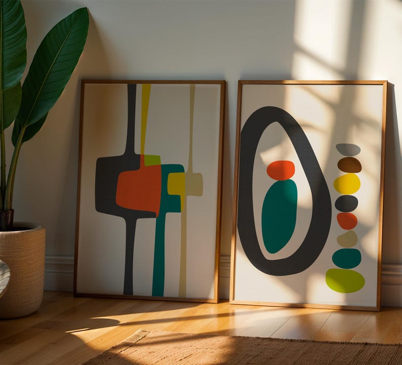 May include: Two framed rectangular wall art prints with light beige backgrounds. The left print features abstract dark gray, orange, teal, and yellow shapes. The right print displays a large dark gray oval enclosing orange and teal shapes, with a vertical stack of multicolored pebbles beside it.