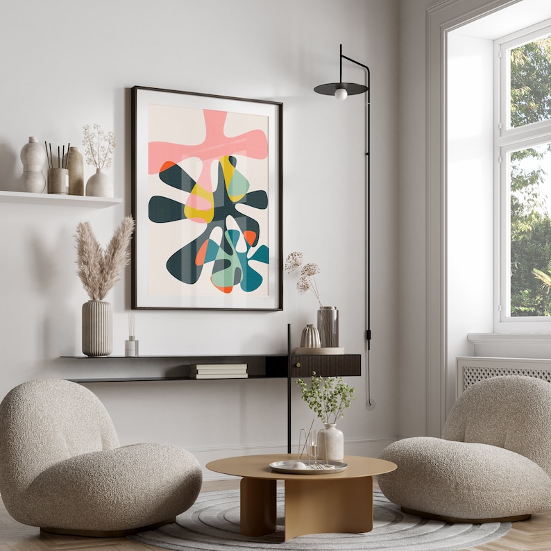 May include: A framed wall art print featuring an abstract composition of organic, amoeba-like shapes in shades of pink, dark teal, yellow, and orange against a light cream background, displayed in a room with two textured beige lounge chairs.