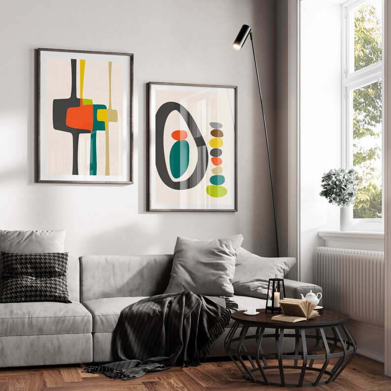 May include: Two framed wall art prints featuring abstract geometric shapes in shades of charcoal, orange, teal, and yellow on a light cream background. The left print displays vertical lines and blocks, while the right print shows a large oval and stacked circles.