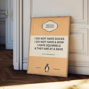 May include: A rectangular framed poster with a light orange background and a central cream-colored horizontal band. The text reads: SPECIAL EDITION, I DO NOT HAVE DUCKS I DO NOT HAVE A ROW I HAVE SQUIRRELS & THEY ARE AT A RAVE, AN AUTOBIOGRAPHY, COMPLETE, UNABRIDGED, with a small black penguin illustration.