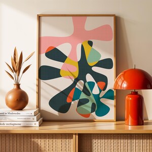 May include: A framed abstract wall art print featuring organic, amoeba-like shapes in pink, dark green, yellow, teal, and orange on a cream background. To the left is a brown vase with dried leaves on books, and to the right is a bright orange dome lamp.