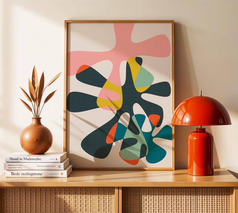 May include: A framed abstract wall art print featuring organic, amoeba-like shapes in pink, dark green, yellow, teal, and orange on a cream background. To the left is a brown vase with dried leaves on books, and to the right is a bright orange dome lamp.