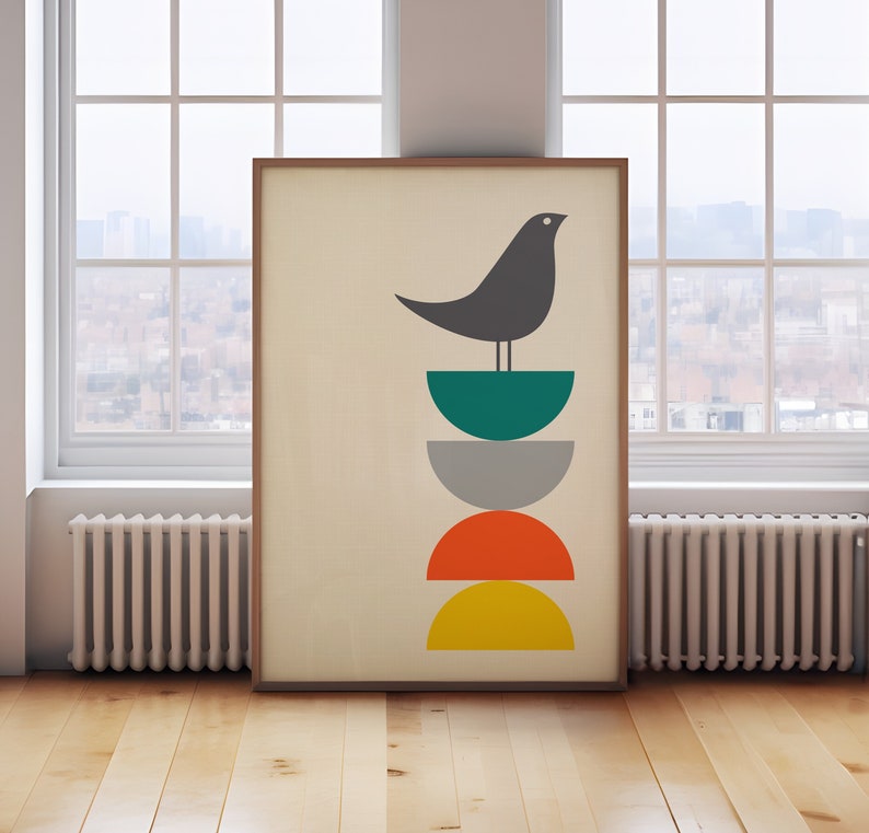 May include: A framed wall art print featuring a dark gray bird perched atop a vertical stack of four semi-circles in teal, gray, orange, and yellow. The design is set against a light cream background, enclosed within a simple wooden frame.
