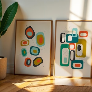 May include: Two framed rectangular wall art prints featuring abstract geometric shapes in shades of orange, teal, olive green, and charcoal gray. The designs include rounded rectangles and thin black lines with small dots at the ends.