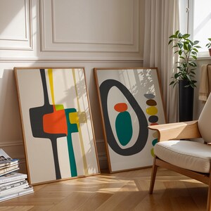 May include: Two rectangular framed wall art prints featuring abstract geometric shapes in black, orange, teal, and yellow on a cream background. The left print displays vertical lines and block forms, while the right print shows a large black oval.