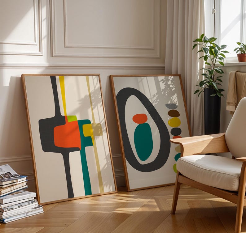May include: Two rectangular framed wall art prints featuring abstract geometric shapes in black, orange, teal, and yellow on a cream background. The left print displays vertical lines and block forms, while the right print shows a large black oval.