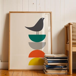 May include: A framed wall art print featuring a dark gray bird perched atop a vertical stack of four semi-circles in teal, light gray, orange, and yellow. The artwork is set against a cream background and enclosed in a light wood frame.