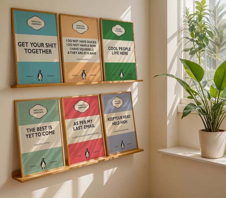 May include: Six framed rectangular wall art prints arranged on two wooden shelves. Each print features a colorful background, a penguin illustration, and text including phrases like GET YOUR SHIT TOGETHER, AS PER MY LAST EMAIL, and COOL PEOPLE LIVE HERE.
