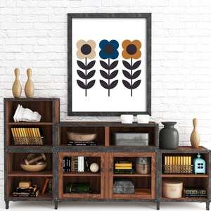 Op de afbeelding: Een ingelijst afdruk van drie gestileerde bloemen met bruine, blauwe en gele bloemblaadjes. De bloemen staan op een witte achtergrond en het frame is zwart. De afdruk staat op een houten plank met andere decoratieve items.