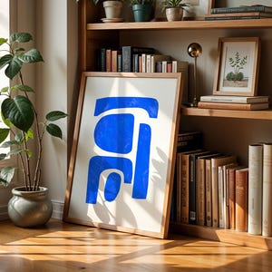 May include: A framed wall art print featuring a bold, abstract blue shape composed of three distinct, rounded geometric segments on a white background, displayed in a light-toned wooden frame resting on a wooden floor.