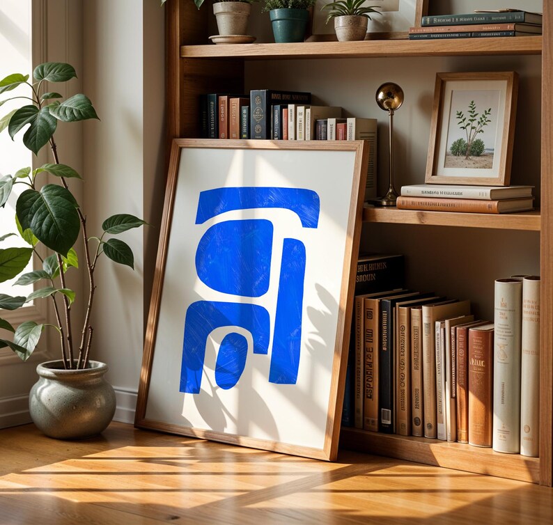 May include: A framed wall art print featuring a bold, abstract blue shape composed of three distinct, rounded geometric segments on a white background, displayed in a light-toned wooden frame resting on a wooden floor.