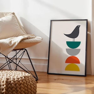 May include: A framed vertical wall art print featuring a dark gray bird perched atop a vertical stack of four semi-circles in teal, gray, orange, and yellow colors. The artwork is set against a light cream background within a thin black frame.