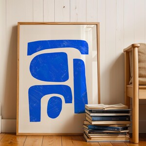 May include: A framed wall art print featuring a series of bold, abstract blue shapes with visible brushstroke textures against a solid off-white background. The artwork is enclosed in a light-toned wooden frame with a clean, simple profile.