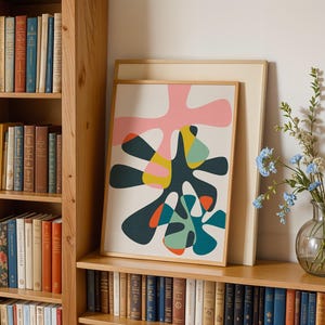 May include: A framed abstract art print featuring organic, amoeba-like shapes in shades of pink, dark teal, yellow, and orange on a cream background. The print is displayed in a light wood frame, positioned on a shelf next to a glass vase of blue flowers.