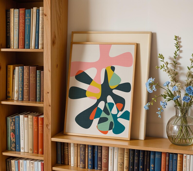 May include: A framed abstract art print featuring organic, amoeba-like shapes in shades of pink, dark teal, yellow, and orange on a cream background. The print is displayed in a light wood frame, positioned on a shelf next to a glass vase of blue flowers.