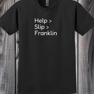 May include: Black t-shirt with white text that reads "Help > Slip > Franklin". The shirt has a crew neck and short sleeves. The text is stacked vertically on the front of the shirt.