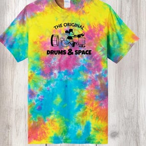 May include: Tie-dye t-shirt with a vibrant mix of yellow, pink, blue, and green. The shirt features a graphic of a cartoon mouse playing drums, with the text "THE ORIGINAL DRUMS & SPACE" in black.