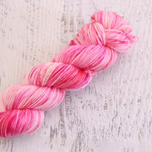 May include: A skein of pink yarn with varying shades of pink and white. The yarn is tightly wound and appears soft, ideal for knitting or crocheting projects. The background is a white, weathered wooden surface.