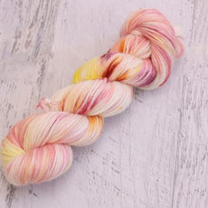 May include: A skein of hand-dyed yarn featuring a variegated color palette of soft cream, pale yellow, and vibrant pink with deep burgundy accents. The yarn is twisted into a traditional hank shape, showcasing a smooth, multi-tonal fiber texture.