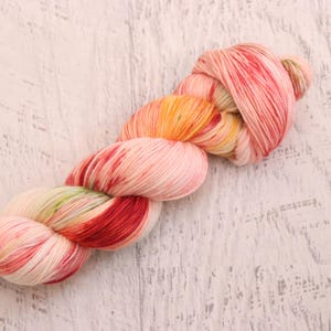 May include: A skein of hand-dyed yarn in a range of colors, including pink, red, orange, and yellow. The yarn is arranged in a twisted bundle, with a soft, textured appearance. The background is a light-colored, weathered wooden surface.