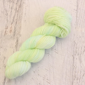 Op de afbeelding: Een streng lichtgroen garen met subtiele kleurvariaties. Het garen is gerangschikt in een gedraaide, langwerpige vorm, die de textuur en kleur laat zien. Het garen ligt op een lichtgekleurd, getextureerd houten oppervlak, dat de kleur van het garen benadrukt.