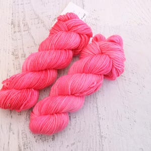 May include: Two skeins of vibrant pink yarn are arranged diagonally on a white, weathered wooden surface. The yarn is tightly wound, showcasing its texture and color. A small tag is visible in the background. This image is ideal for craft projects.