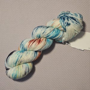 May include: A skein of hand-dyed yarn in a white base with splashes of blue, teal, orange, and red. The yarn is twisted into a loose coil and has a tag attached.