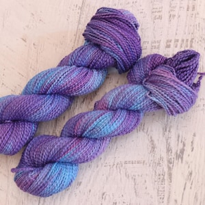 May include: Two skeins of hand-dyed yarn in shades of purple, blue, and pink. The yarn is twisted into a rope-like shape, with a textured appearance. The background is a light-coloured, weathered wooden surface.