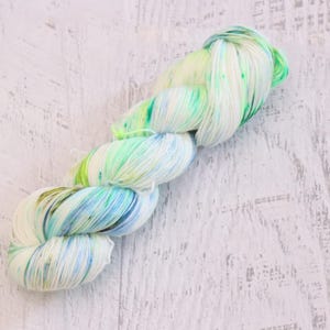 May include: A twisted skein of yarn featuring a variegated color palette of white, bright lime green, and soft sky blue. The fibers are tightly wound into a long, braided structure, showcasing a blend of cool tones and vibrant accents throughout.