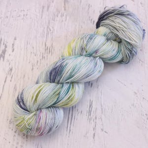 May include: A single skein of hand-dyed yarn featuring a variegated color palette of soft light blue, pale lime green, and deep indigo speckles. The twisted fiber bundle displays a multi-tonal appearance with fine strands of twisted wool.