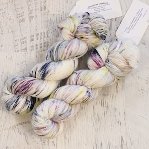 May include: Two skeins of speckled yarn in cream, purple, navy, and yellow. The yarn is labeled "ChemKnits Creations" with details about the fiber content and weight. The yarn is on a white, distressed wooden surface.