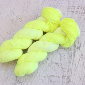 May include: A skein of vibrant neon yellow yarn, suitable for knitting or crochet. The yarn is tightly wound and has a soft, fluffy texture. The background is a light-coloured, distressed wood surface.