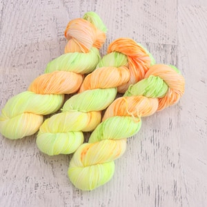 May include: Three skeins of hand-dyed yarn twisted into bundles. The yarn features a variegated color palette of bright neon yellow, soft lime green, and vibrant orange, with the colors blending together throughout the length of each fiber strand.