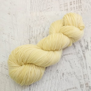 May include: A skein of pale yellow yarn, twisted into a figure eight shape, against a white wood background.