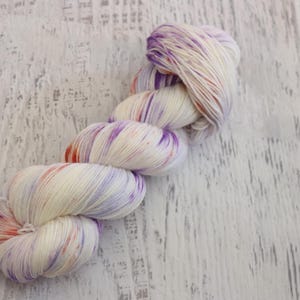 May include: A skein of white yarn with purple, orange, and pink speckles. The yarn is twisted into a loose coil.