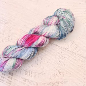 May include: A twisted skein of yarn featuring a variegated color palette of bright magenta, soft light blue, and white. The fibers are tightly wound into a long, cylindrical shape with visible individual strands throughout the entire length.