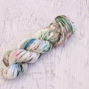May include: A twisted skein of yarn featuring a cream-colored base with variegated speckles and streaks of forest green, sky blue, and soft pink. The yarn is wound into a long, continuous loop with a textured, multi-colored appearance throughout.