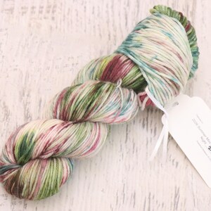 May include: A skein of hand-dyed yarn featuring a variegated color palette of cream, sage green, deep forest green, and muted burgundy. A white rectangular paper tag is attached to the yarn, printed with the text 2024 Summer Mini Skein Mini Series Night 2.