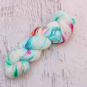 May include: A skein of hand-dyed yarn in a variegated colorway. The yarn features a blend of white, turquoise, pink, and red hues. The yarn is arranged in a twisted, braided form, showcasing the color variations. The background is a white, textured surface.