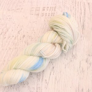May include: A twisted skein of yarn featuring a soft color palette of cream, pale yellow, and light blue. The fibers are tightly wound into a long, cylindrical shape, showcasing a variegated pattern of pastel hues throughout the entire length.