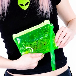 May include: A black t-shirt with a green alien head graphic and a neon green, clear, zippered pouch filled with dark green glitter. The pouch has a matching wrist strap. The person is wearing a black denim skirt.