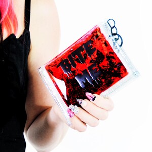 May include: A rectangular clear plastic pouch filled with a red liquid and black glitter, featuring the text BITE ME in bold black letters. The pouch has a small black loop attached to the top corner and a clear, sealed edge design.