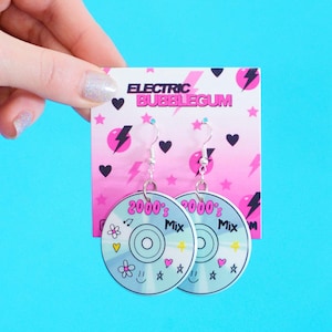 May include: A pair of dangle earrings featuring a CD design with the text "2000's Mix". The earrings are held against a pink and white card with the text "Electric Bubblegum". The earrings have silver hooks and blue accents.