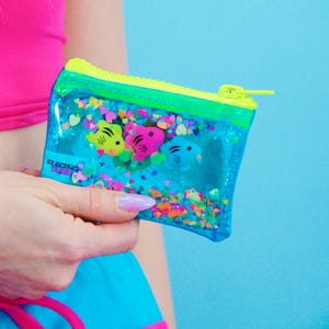 May include: A clear, rectangular pouch with a neon yellow zipper and trim. The pouch contains colorful confetti and three fish-shaped charms in yellow, pink, and blue. The text "ELECTRIC BUBBLE" is visible on the pouch.
