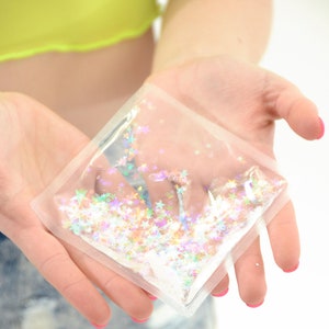 May include: A clear, square-shaped pouch filled with colorful star-shaped confetti. The confetti includes shades of pink, purple, green, and white. The pouch is held in open hands against a light background.