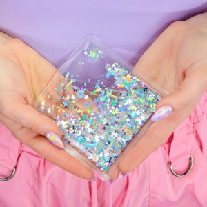 May include: A clear plastic pouch filled with iridescent glitter in the shape of stars and crescent moons. The pouch is held in two hands.