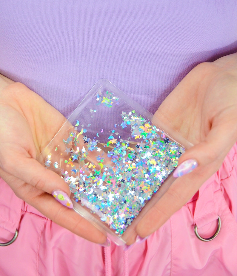 May include: A clear plastic pouch filled with iridescent glitter in the shape of stars and crescent moons. The pouch is held in two hands.